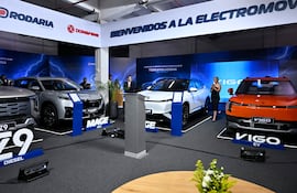 Mujer de negro interactúa con la audiencia en un evento de electromovilidad, con varios vehículos eléctricos exhibidos a su alrededor.