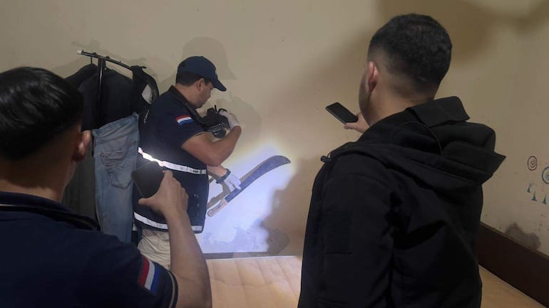 Hombre en uniforme con guantes examina un objeto iluminado, mientras otro documenta desde un teléfono en una habitación desordenada.