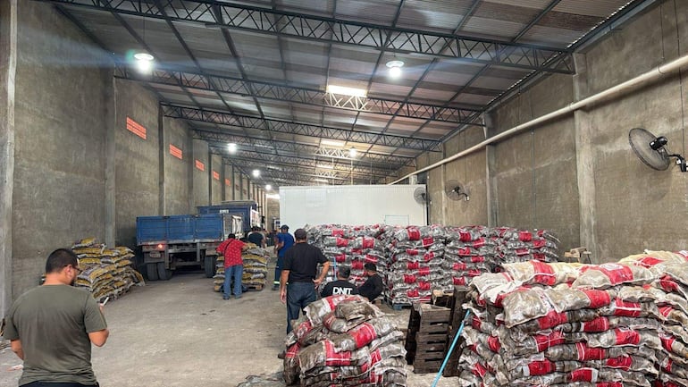 Seis hombres en un galpón, uno usa un teléfono móvil mientras organizan sacos rojos en un ambiente iluminado artificialmente.