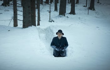 Hombre en abrigo oscuro y sombrero negro, parcialmente enterrado en nieve, observando hacia la cámara en un bosque nevado.