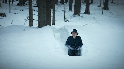 Hombre en abrigo oscuro y sombrero negro, parcialmente enterrado en nieve, observando hacia la cámara en un bosque nevado.