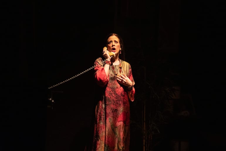 Mujer vestida con colores rojos y flores, hablando seriamente por teléfono con mano sobre el pecho, en ambiente teatral iluminado.