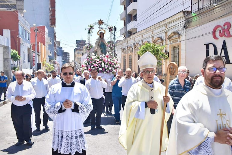 Sacerdote con báculo y túnica blanca lidera a la multitud, hombres con camisas blancas portan un altar decorado con flores.