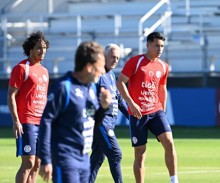 Cuatro hombres en campo de entrenamiento, tres en camisetas rojas con logo de Tigo y un asistente con equipo oscuro, observando.