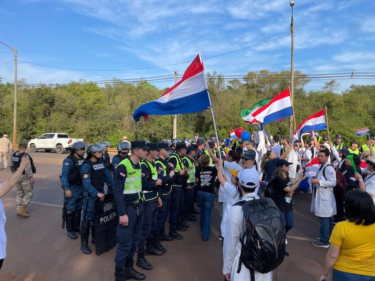 Grupo de policías alineados frente a manifestantes con batas blancas y banderas, en un ambiente tenso y de protesta.