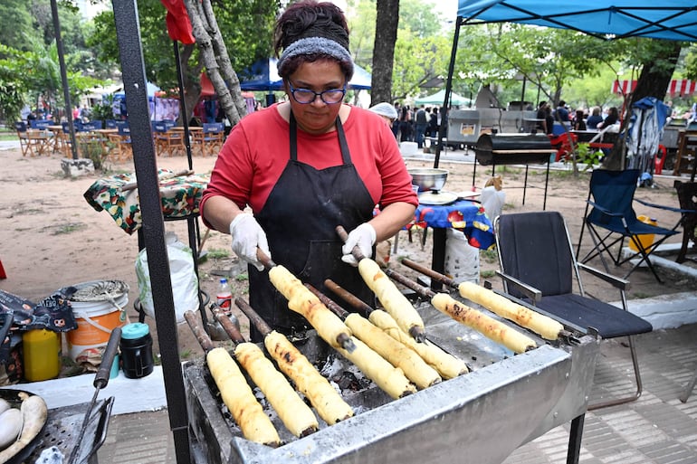 Mujer de cabello recogido con suéter rojo y delantal negro cocina a la parrilla en un ambiente festivo rodeada de árboles.
