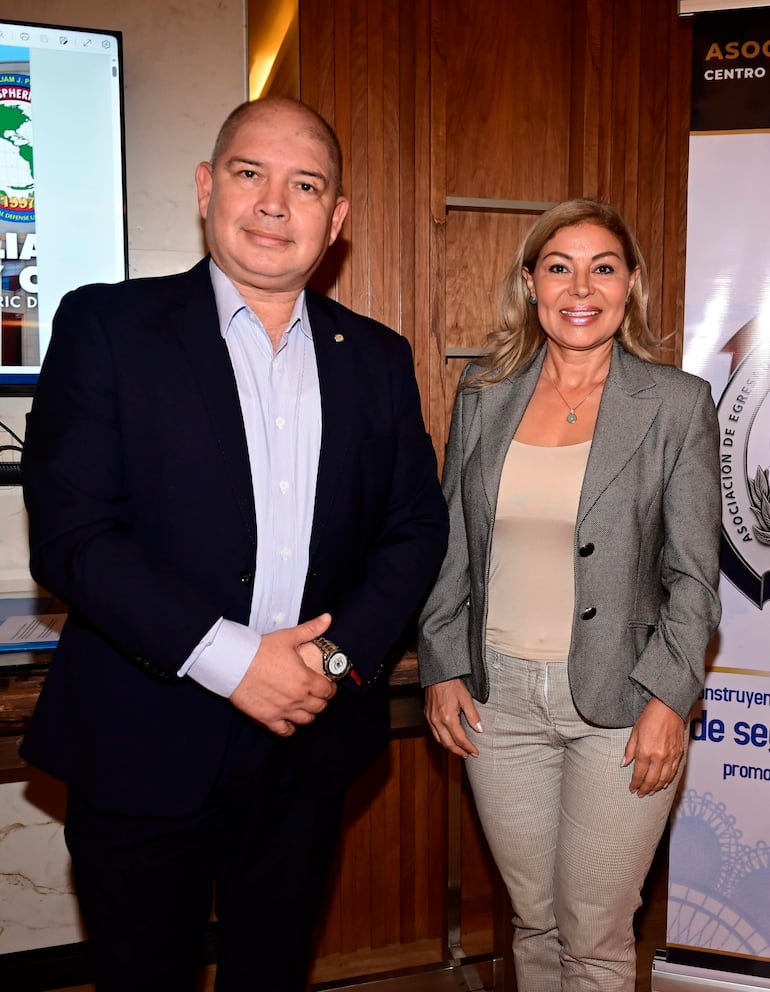 Hombre con traje oscuro y camisa clara y mujer con chaqueta gris sonríen en evento de egresados en ambiente acogedor.