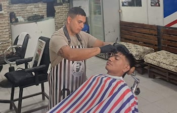 Barbero tatuado con guantes corta el cabello de un cliente con capa roja, blanca y azul, en barbería decorada con ladrillos expuestos.