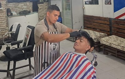 Barbero tatuado con guantes corta el cabello de un cliente con capa roja, blanca y azul, en barbería decorada con ladrillos expuestos.