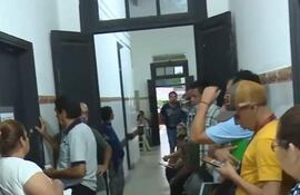 Grupo de personas de diversas edades en pasillo hospitalario, conversando y mirando dispositivos móviles, con ambiente interno y simple decoración.
