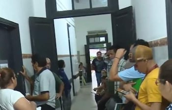 Grupo de personas de diversas edades en pasillo hospitalario, conversando y mirando dispositivos móviles, con ambiente interno y simple decoración.