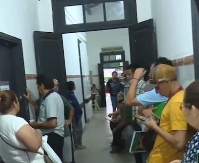 Grupo de personas de diversas edades en pasillo hospitalario, conversando y mirando dispositivos móviles, con ambiente interno y simple decoración.