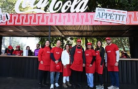 Seis personas sonríen en delantales y gorras rojas de Coca-Cola, posando en un evento al aire libre con vegetación de fondoAndrea Aponte, Alejandro Nahmias, Alejandra Gómez, Luciano Weston, Adriana Acosta, Macarena Pascottini,
y Fabrizzio Rehnfeldt.s asisten a un evento gastronómico al aire libre promovido por Coca-Cola.