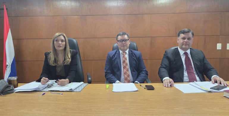 Inés Galarza con cabello rubio y saco negro sostiene una carpeta, Pablino Barreto en traje azul y Juan Dávalos en traje negro observan atentos.