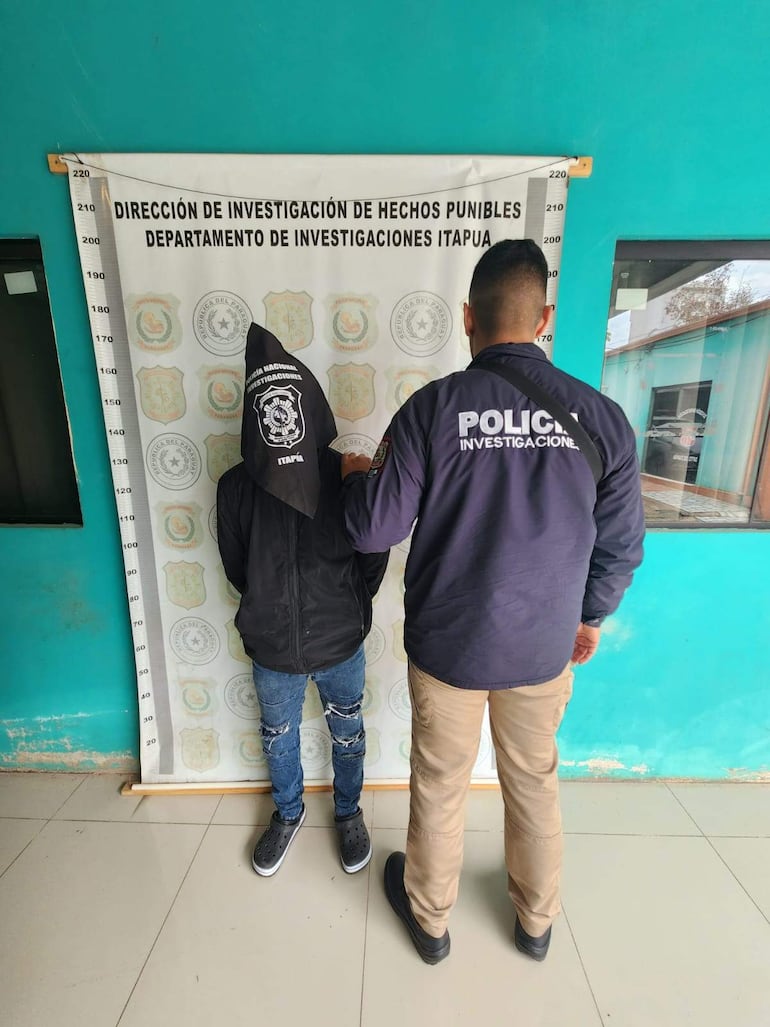 Oficial de policía con chaqueta 'POLICÍA INVESTIGATIVA' custodia a un hombre con capucha y chaqueta negra en oficina de investigaciones.