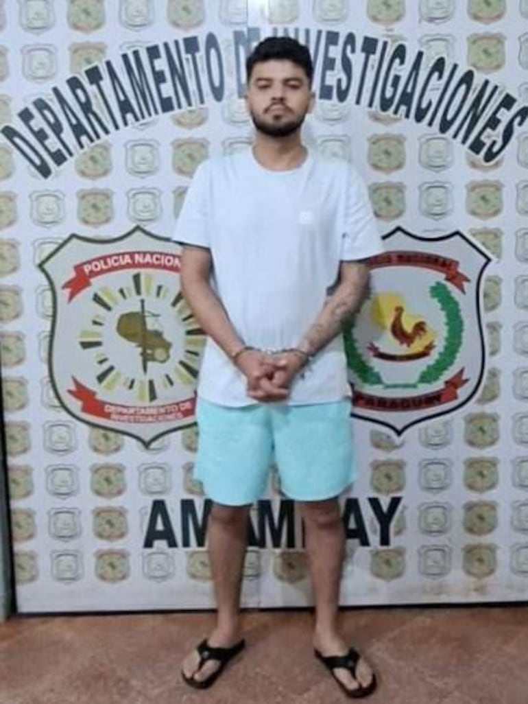 Hombre de pie con camiseta blanca y pantalones cortos celestes, manos atadas, frente a un mural de la Policía Nacional de Paraguay.
