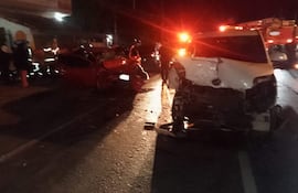 Escena de accidente automovilístico con vehículos destrozados y bomberos atendiendo a los heridos en la oscuridad.