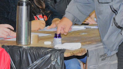 Manos señalando frasco de tinta morada y sosteniendo un bolígrafo, sobre mesa cubierta con tela negra.