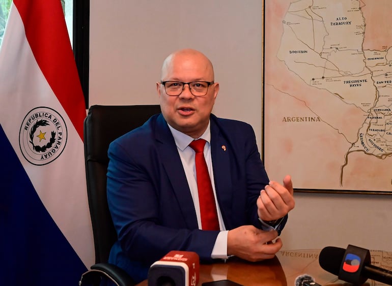 Óscar Lovera en traje azul y corbata roja gesticula mientras habla, con bandera paraguaya y micrófonos en la mesa.