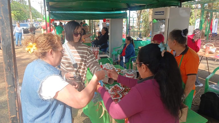 Cuatro mujeres participan en un intercambio de productos en un evento al aire libre decorado con globos y carpas verdes.