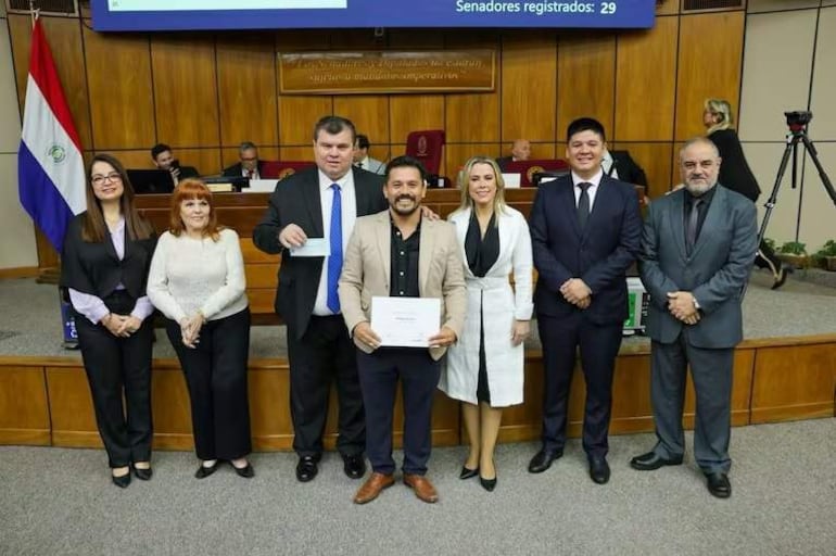 Seis personas vestidas formalmente, dos sosteniendo documentos en un ambiente institucional con banderas paraguayas de fondo.