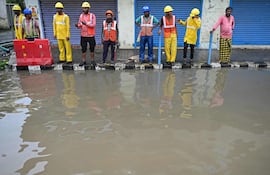 Hombres de pie junto a una acera a lo largo de una calle inundada después de las lluvias, mientras se espera que el ciclón Michaung toque tierra.
