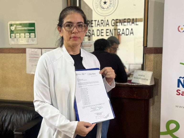 Mujer con chaqueta blanca sostiene un documento mientras en el fondo se observa una mesa con otras personas en un ambiente administrativo.