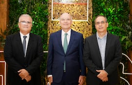 Tres hombres de pie en trajes formales. Manuel Fronciani al centro sonriente, rodeado de hombres con gafas y sacos oscuros.