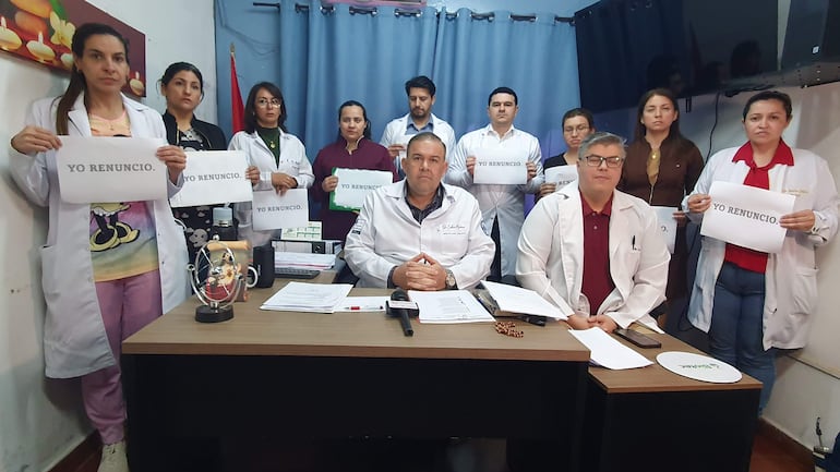 Grupo de médicos en bata blanca sosteniendo carteles que dicen 'YO RENUNCIO', con expresión seria y ambiente de protesta.