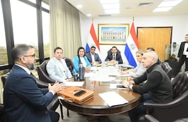 Latorre Martínez en traje conversando mientras Centurión González y Meza lo observan. Fondo con banderas de Paraguay y documentos sobre la mesa.