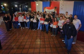 Cincuenta personas con diplomas en un salón decorado en rojo y blanco, expresando felicidad durante una ceremonia de reconocimiento.