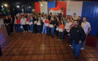 Cincuenta personas con diplomas en un salón decorado en rojo y blanco, expresando felicidad durante una ceremonia de reconocimiento.