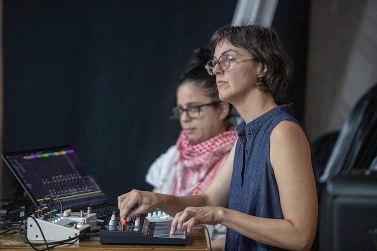 Mujer con gafas y pañuelo rojo en fondo, otra con camisa azul manipulando equipo musical en un entorno creativo.