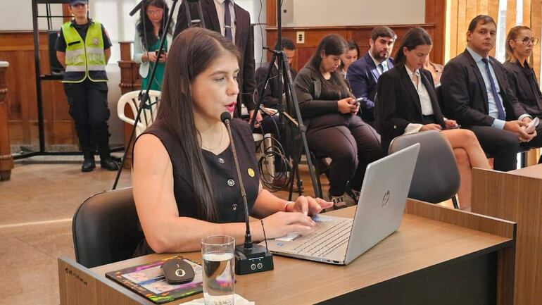Cinthia Vera, vestida de blusa negra, habla frente a una laptop, con audiencia y un miembro de seguridad presente.