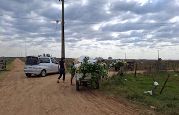Una mujer con camiseta oscura camina mientras un hombre carga materiales verdes en un carrito, en un camino de tierra rural.