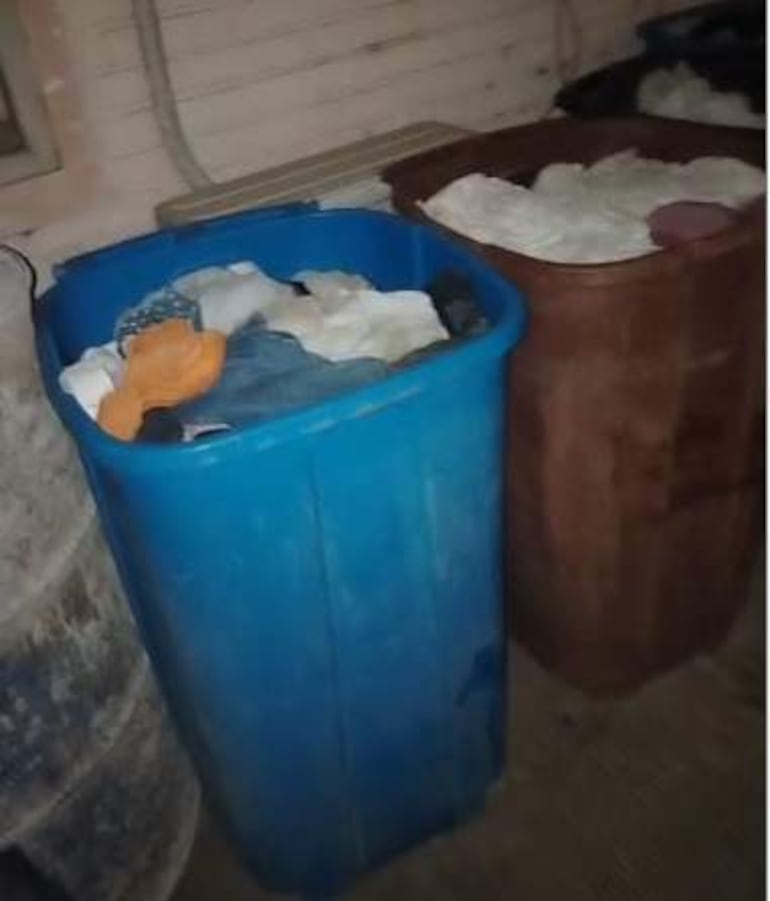 Contenedores grandes llenos de ropa en lavandería, piso gris. Ambiente de trabajo desordenado y precario.