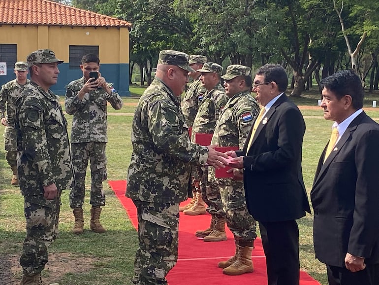 Soldados alineados en uniforme camuflado observan a un hombre en traje negro y corbata amarilla mientras recibe un reconocimiento.