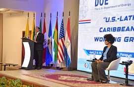 Félix Sosa en podio con traje oscuro, mientras mujer con auriculares lo escucha atentamente, rodeados de banderas de países americanos.