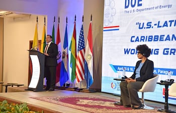 Félix Sosa en podio con traje oscuro, mientras mujer con auriculares lo escucha atentamente, rodeados de banderas de países americanos.