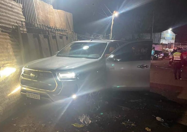 Toyota Hilux plateada con puerta abierta y faros encendidos, en un entorno oscuro y descuidado, sin personas visibles.