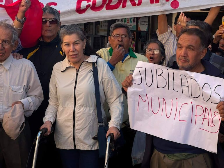 Mujer con andador y hombre mayor con gafas lideran protesta de jubilados, mientras otros sostienen carteles en un ambiente urbano.