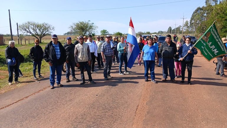 Pobladores sosteniendo una bandera paraguaya y un cartel, reunidos en carretera rural con expresiones tranquilas y algunas preocupadas.