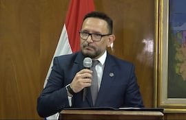 Derlis León, en traje oscuro y corbata clara, habla al micrófono frente a un fondo de madera y una bandera paraguaya.