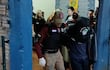 Agente en chaleco táctico sostiene a un hombre cabizbajo en un entorno cerrado, reflejando una situación de control de seguridad.