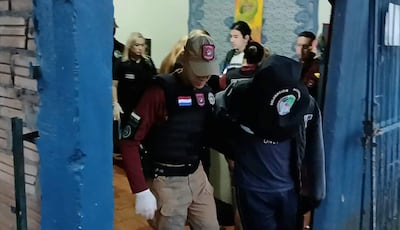 Agente en chaleco táctico sostiene a un hombre cabizbajo en un entorno cerrado, reflejando una situación de control de seguridad.