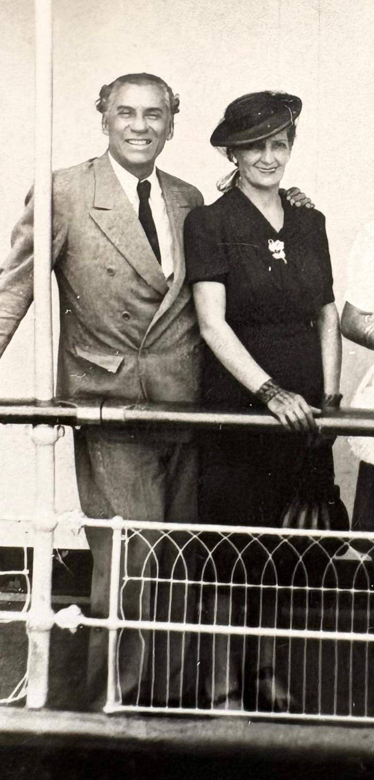 Hombre canoso en traje claro y mujer en atuendo negro con sombrero, ambos sonríen en la cubierta de un barco.
