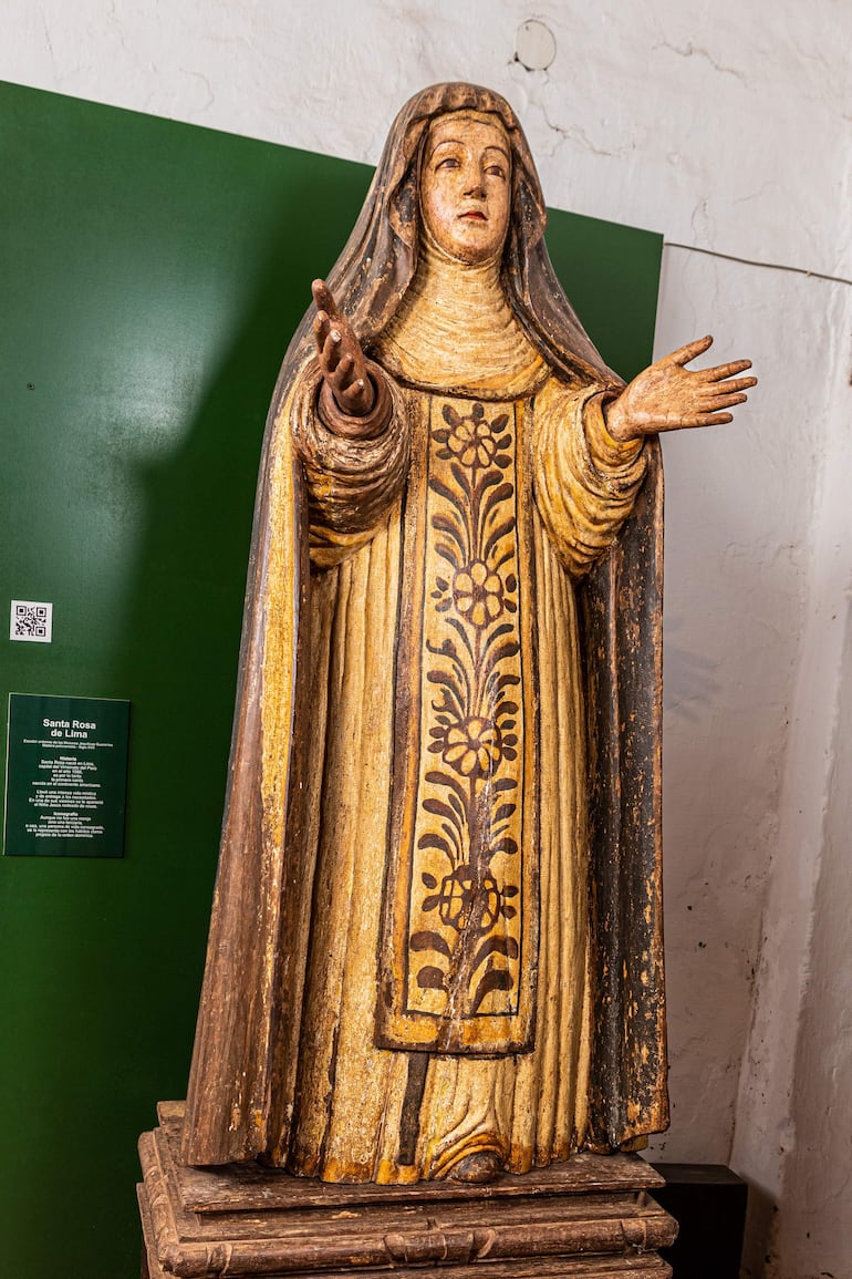 Estatua de Santa Rosa de Lima en madera, vestida con diseño floral y expresión serena, se exhibe frente a fondo verde.