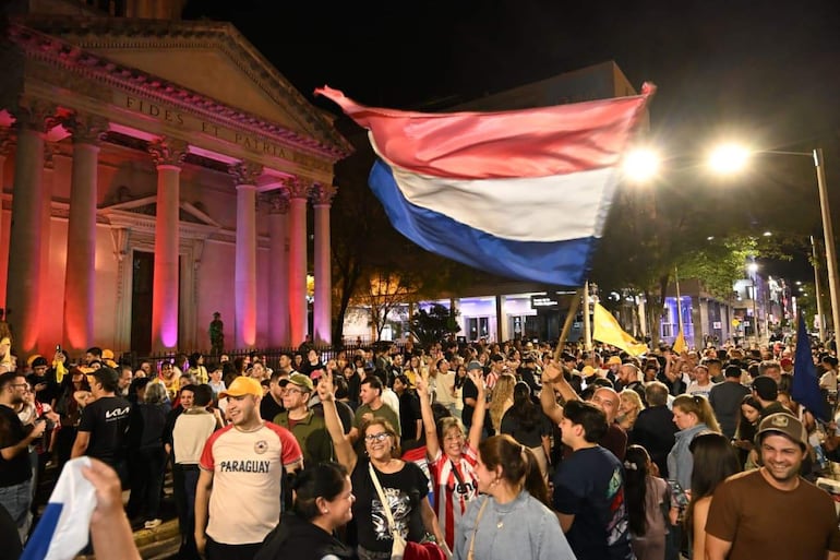 Multitud sonriente sostiene una gran bandera paraguaya, vestida con camisetas de colores en un ambiente festivo y luminoso.
