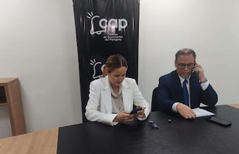 Soledad Núñez en vestido blanco revisando su teléfono, mientras Gustavo Egüez en traje azul habla por teléfono en la mesa.
