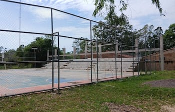 Cancha de baloncesto al aire libre en desuso, con gradas de concreto y vegetación alrededor en un día nublado.